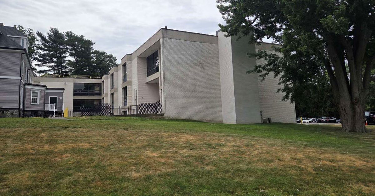 A three-story pale brick building with recessed balconies behind a lawn and a large tree, next to a gray clapboard building.
